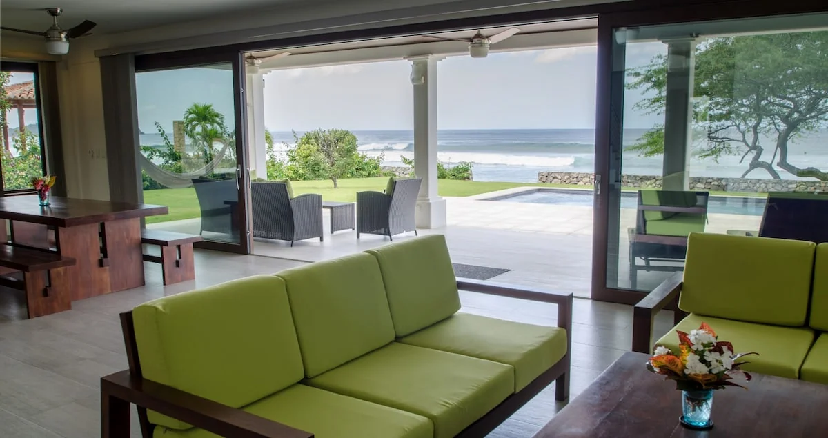 A bright living room with lime-green seating opens through glass doors to a patio, lawn, and ocean beyond.