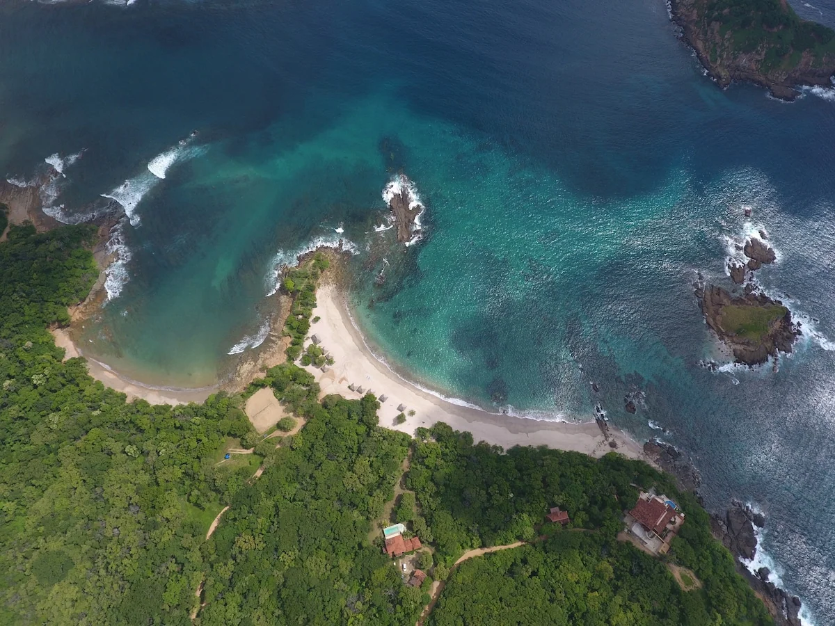 An aerial view shows a curved sandy beach, turquoise ocean water, rocky outcrops, and dense green forest with a few buildings.