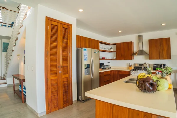 A bright kitchen with wooden cabinets, a stainless-steel refrigerator, a large island, and a fruit basket beside a staircase.