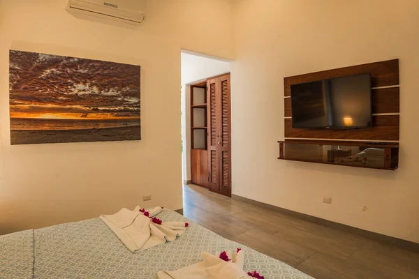 A warmly lit bedroom contains a made bed with decorative towels, a wall-mounted television, a landscape painting, and an open doorway.