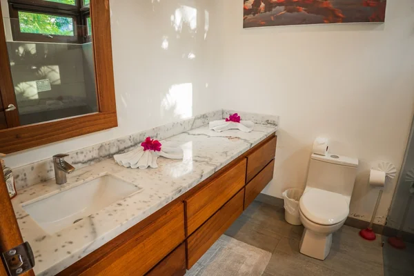 A bright bathroom with a marble vanity, wooden cabinets, a toilet, and small pink flower decorations.