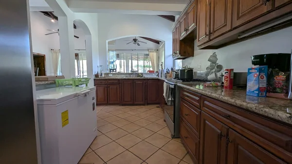 A spacious kitchen with dark wood cabinetry, tiled flooring, granite countertops, a refrigerator, and a large chest freezer.
