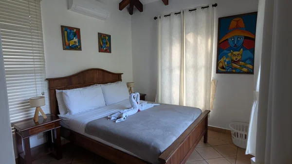 A tidy bedroom with a wooden bed, gray bedding, white curtains, framed colorful artwork, and bedside lamps.