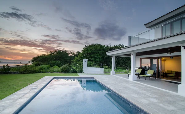 A rectangular swimming pool beside a modern white house opens onto a tiled patio, lawn, trees, and a sunset sky.