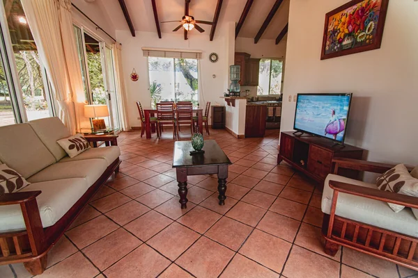 A warmly furnished living room with tiled flooring, seating, a television, dining area, and large windows.