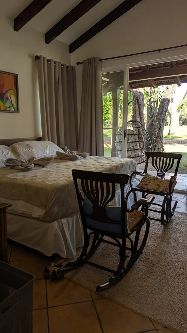 A warmly lit room contains a bed, rocking chairs, a small table, curtains, and large open windows overlooking greenery.