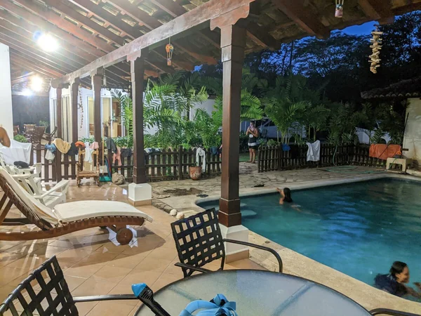 A covered patio with lounge chairs and a table overlooks a lit swimming pool surrounded by plants, with several people in and around the water.