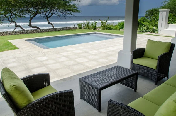 A bright patio with green-cushioned wicker seating overlooks a rectangular pool, landscaped grounds, and the ocean.
