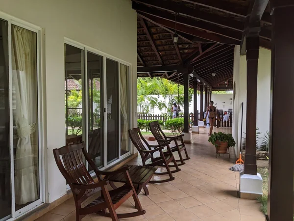 A covered tiled terrace with wooden rocking chairs, large windows, potted plants, and two people in the background.