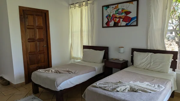 A bright bedroom contains two single beds with white linens, wooden nightstands, a framed colorful painting, and a wooden door.