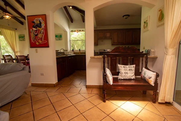 A warmly lit interior shows a tiled living area with a cushioned wooden bench, sofa, dining chairs, artwork, and an adjoining kitchen.