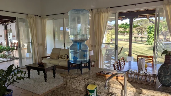 A bright open kitchen and sitting area with a large water dispenser, seating, plants, and windows overlooking a garden.