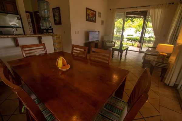 A warmly furnished dining area with a large wooden table, chairs, a television, and glass doors opening toward greenery.