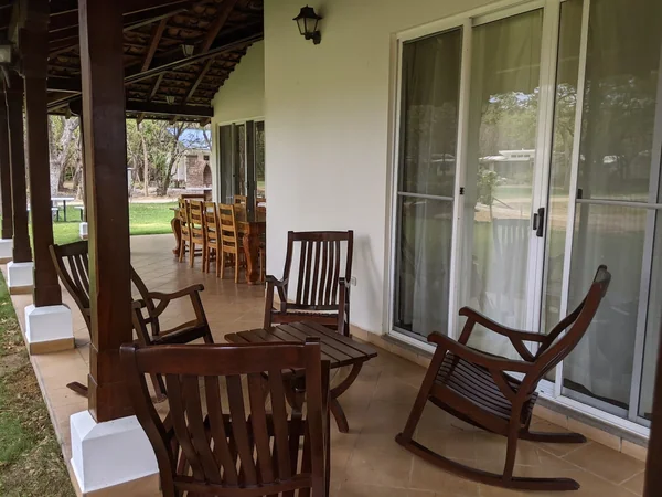 A covered terrace features several wooden rocking chairs, a small table, sliding glass doors, and a dining set extending toward the garden.