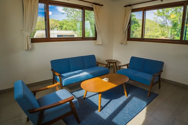 A bright living room with blue seating, wooden tables, a blue rug, and large windows overlooking greenery.