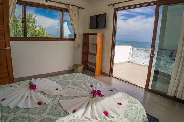 A bedroom with floral towel decorations on the bed, a wall-mounted television, and an open balcony overlooking the ocean.