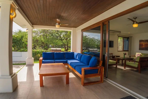 A covered terrace has a blue sectional sofa, wooden coffee table, ceiling fans, and open sliding doors leading into an interior seating area.
