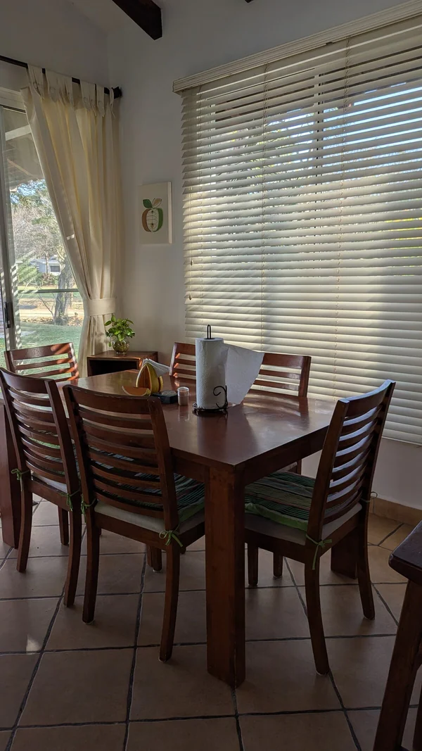 A wooden dining table surrounded by chairs sits beside large windows with blinds and curtains.