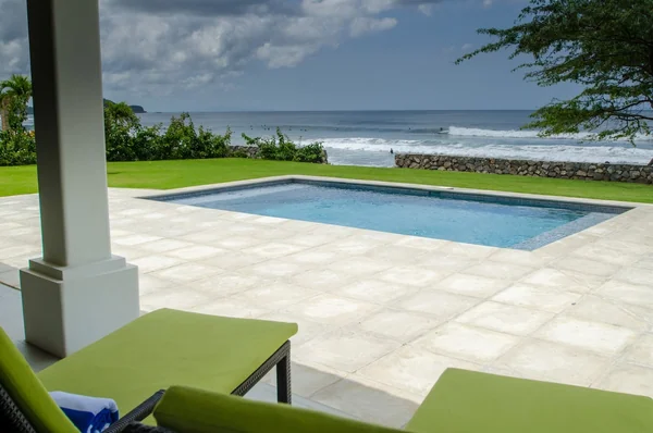 A tiled pool patio with green seating overlooks a rocky shoreline and ocean under a partly cloudy sky.