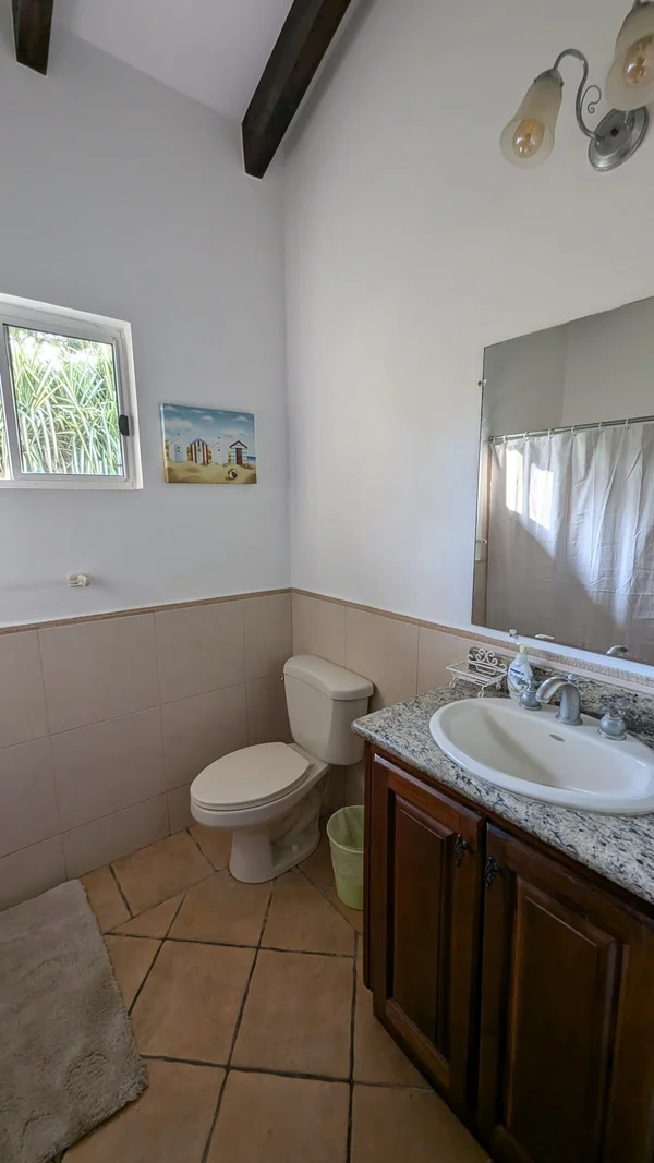 A bright bathroom contains a toilet, pedestal-style sink, wooden vanity, mirror, curtained window, and tiled walls and floor.