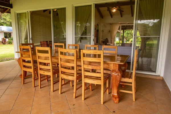 A large wooden dining table surrounded by chairs sits on a tiled covered patio beside glass doors and windows.