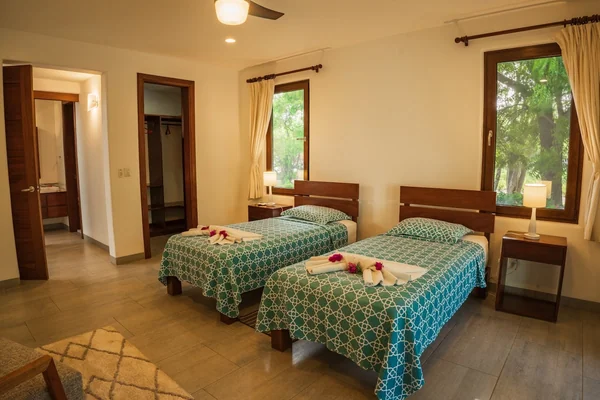 A warmly lit bedroom contains two single beds with patterned teal bedspreads, bedside tables, lamps, windows, and an open wardrobe.