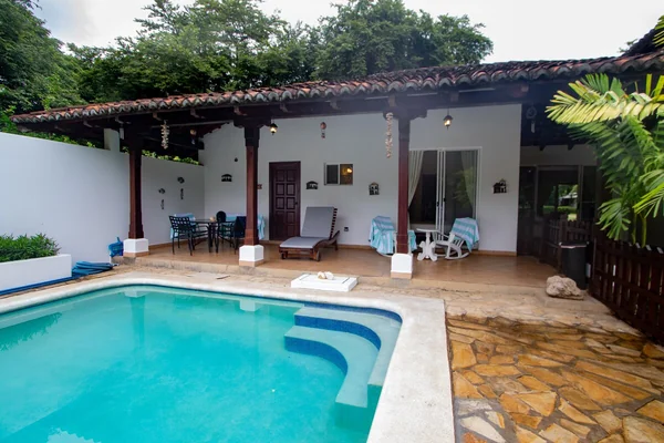 A turquoise swimming pool sits beside a covered patio with seating, loungers, and a white-walled house.