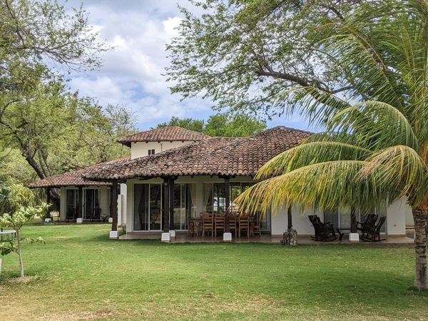 A tiled-roof house with glass walls, outdoor seating, and a palm tree overlooking a broad lawn.