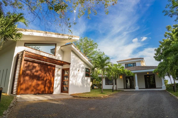 A modern white house with a large wooden garage door, driveway, palm trees, and a bright blue sky.