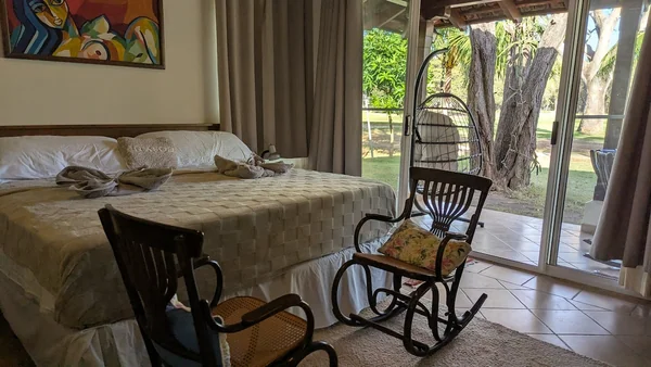 A bedroom contains a large bed, two rocking chairs, a colorful wall painting, and glass doors opening toward a wooded outdoor area.