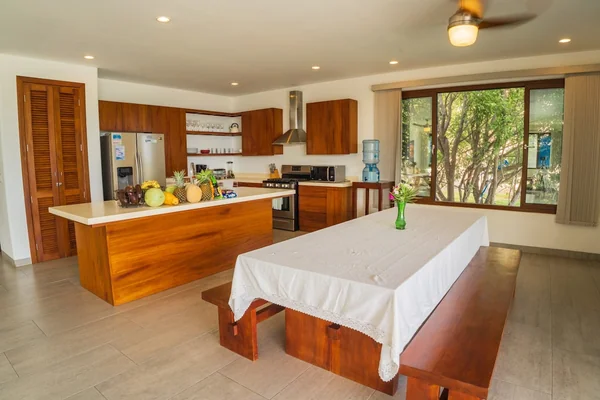 A bright open kitchen and dining area with a wood island, a long table covered in white cloth, fruit, and a large window overlooking trees.