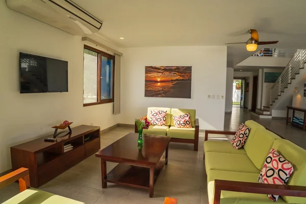 A bright living room contains green sofas with patterned cushions, a wooden coffee table, a wall-mounted television, artwork, and a staircase.