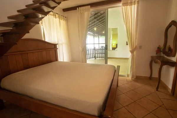 A warmly furnished bedroom with a large wooden bed, exposed staircase, cream curtains, tiled floor, and a balcony doorway.