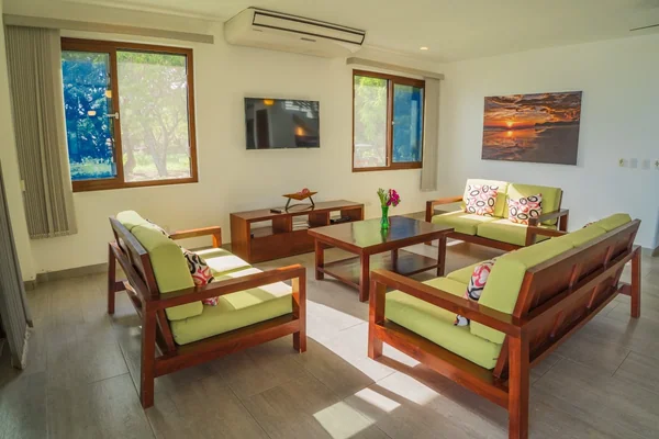 A bright living room contains green-cushioned wooden seating, a glass coffee table, a wall-mounted television, windows, and a landscape painting.