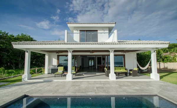 A bright white two-story house with a covered patio, outdoor seating, hammock, garden, and swimming pool.
