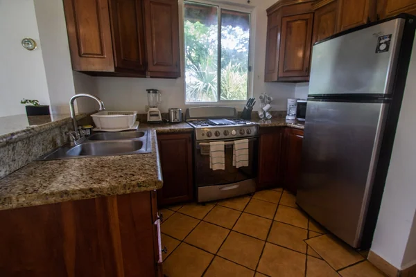 A warm-toned kitchen with wooden cabinets, granite countertops, a sink, stove, refrigerator, and a window overlooking greenery.