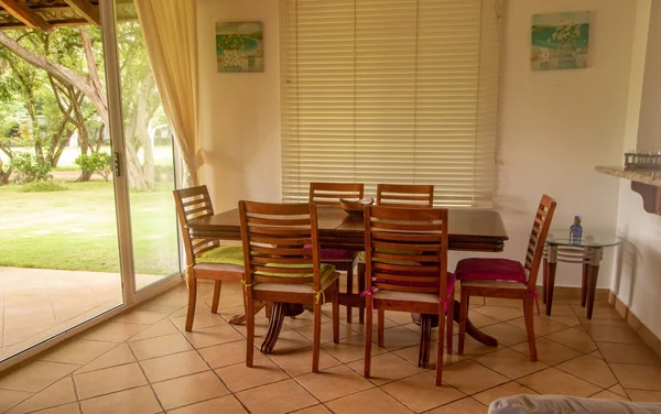 A wooden dining table surrounded by chairs sits beside large glass doors overlooking a green lawn.