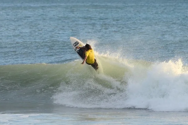 A surfer in a yellow shirt rides a breaking ocean wave while holding a surfboard.