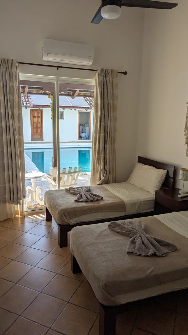 A bright bedroom with two beds, patterned curtains, a ceiling fan, and a view of a pool through the glass doors.