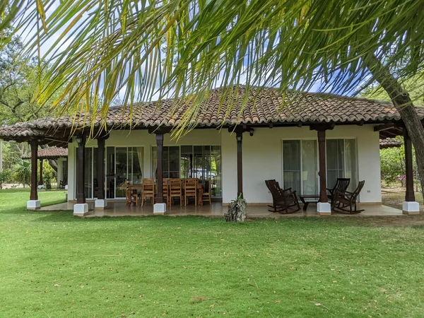 A single-story house with a tiled roof, covered patio, outdoor furniture, and a broad green lawn beneath palm fronds.