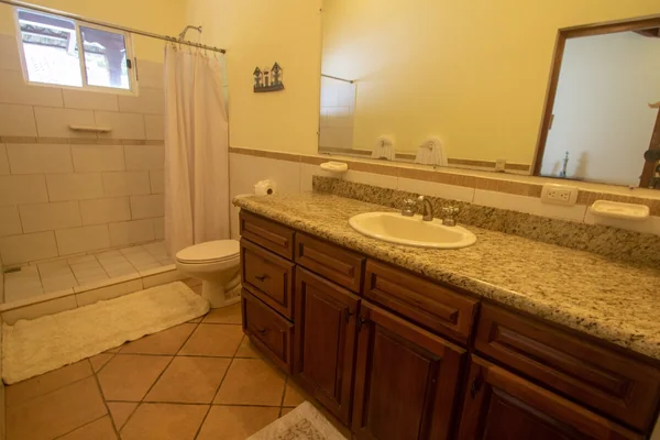 A warmly lit bathroom with a tiled bathtub area, toilet, granite vanity, sink, and large mirror.