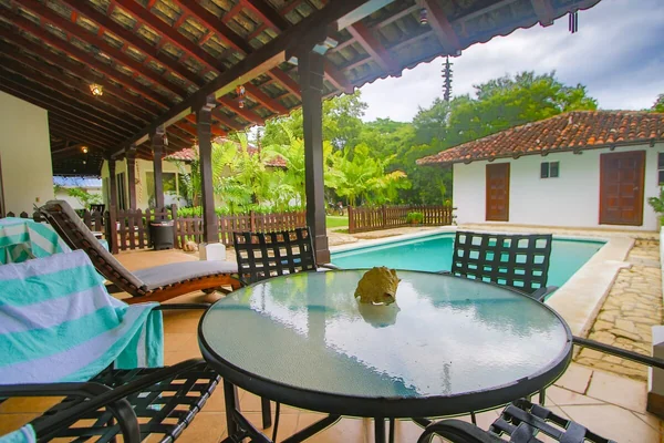 A covered patio with a round glass table, lounge chairs, a swimming pool, and a small white-tiled-roof building surrounded by greenery.