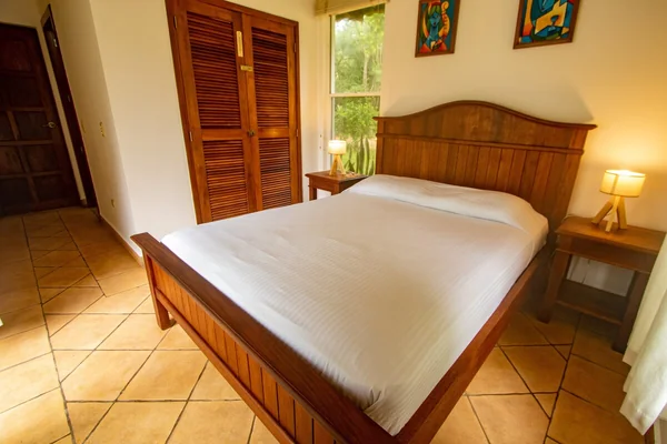 A neatly made wooden bed occupies a warmly lit bedroom with tiled flooring, bedside lamps, a louvered wardrobe, a window, and wall art.
