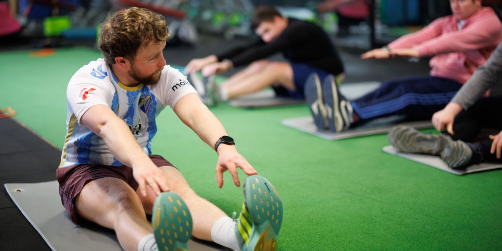 A group class in a gym with participants siitng on the floor doing leg stretch.