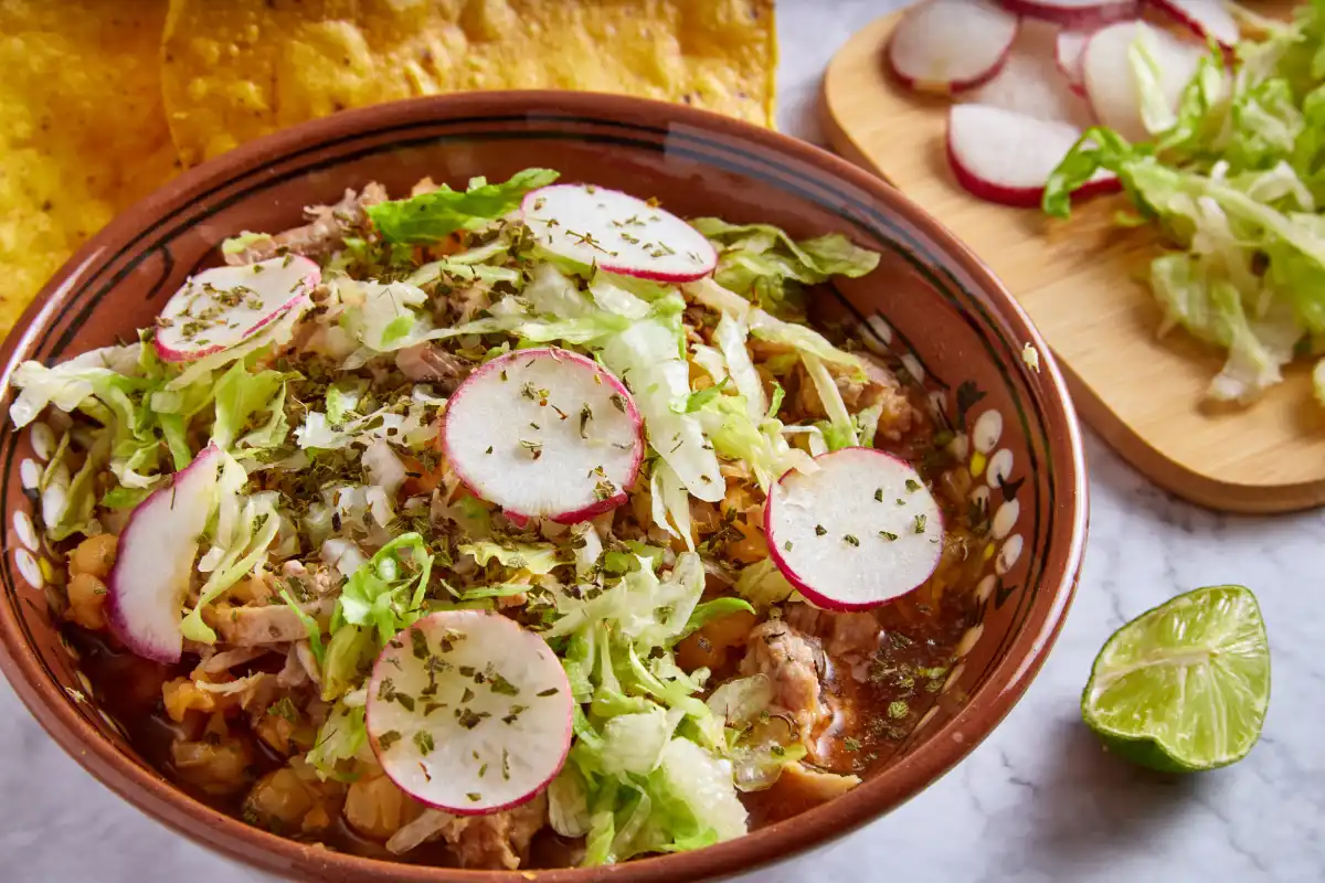 Plato con pozole rojo con rábanos, lechuga y orégano 