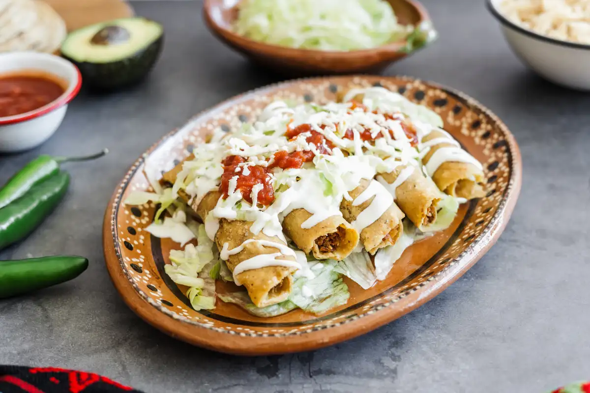 Tacos dorados con lechuga, queso, crema, y salsa roja