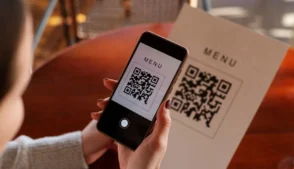 Pessoa usando um celular para escanear um QR Code de um menu impresso sobre uma mesa.