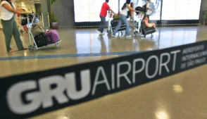 Passageiros empurram carrinhos de bagagem em área interna do aeroporto de Guarulhos, com painel de informações ao fundo.