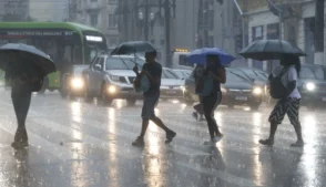 Pedestres atravessam uma rua sob chuva intensa em São Paulo, usando guarda-chuvas, enquanto carros e um ônibus circulam pela via molhada.