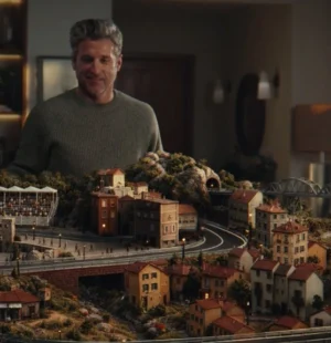 Patrick Dempsey aparece diante de uma pista de autorama em miniatura, cercada por casas, montanhas e arquibancadas, em cena da campanha do Nubank Ultravioleta.